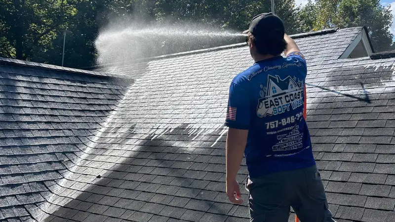 A worker in a blue shirt pressure washes a dark-colored shingle roof, with water mist spraying into the air on a sunny day.