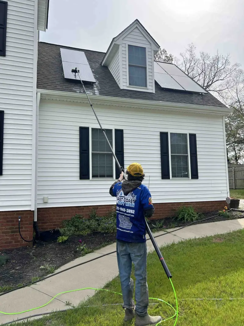 A worker in a blue shirt and yellow hat uses a long pole to clean solar panels on a house’s sloped roof.