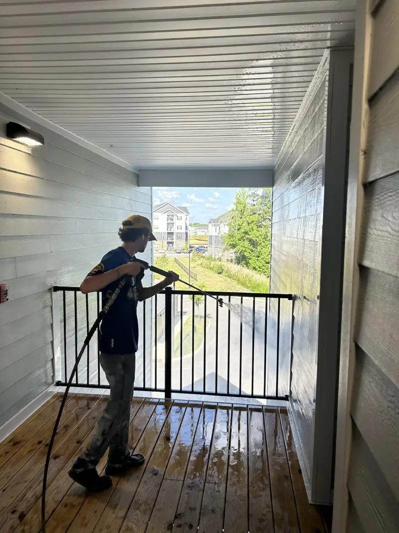 A worker in a yellow hat pressure washes a wooden balcony with black railings, with water spraying towards the railing.