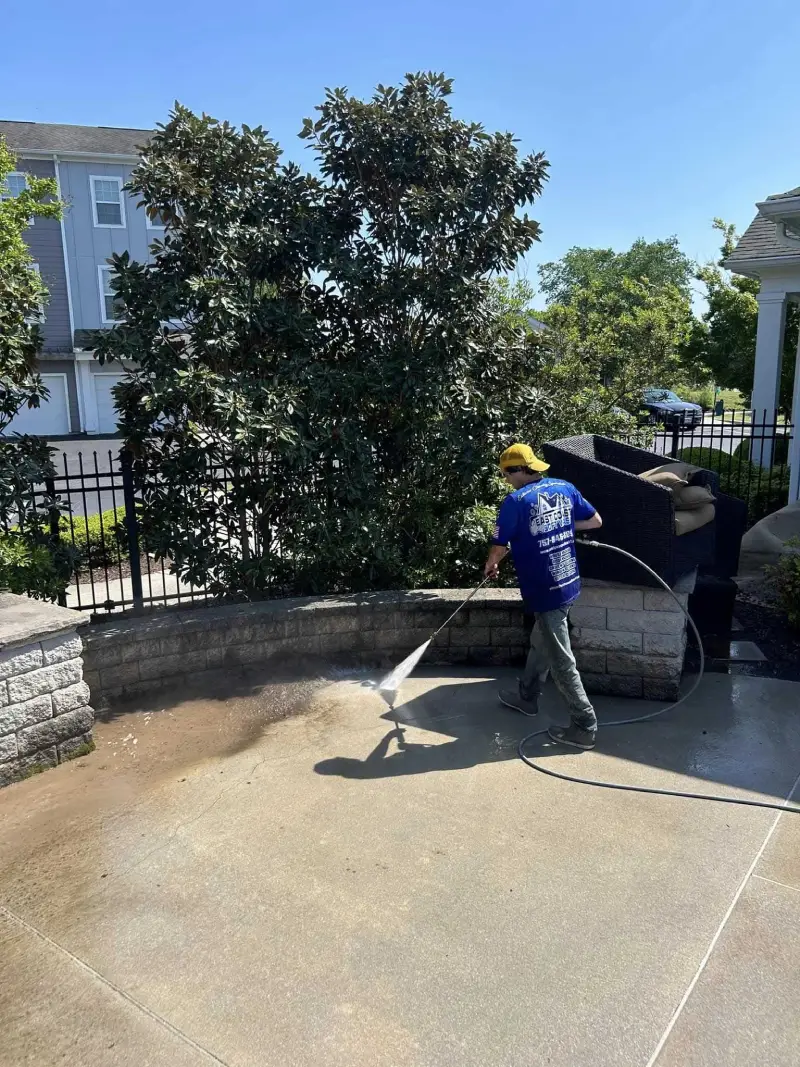 A worker pressure washing the concrete pavement near a seating area with black outdoor furniture and greenery.