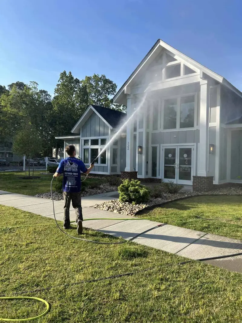 A worker in a blue uniform using a pressure washer to clean an outdoor concrete patio near a fence and some plants.